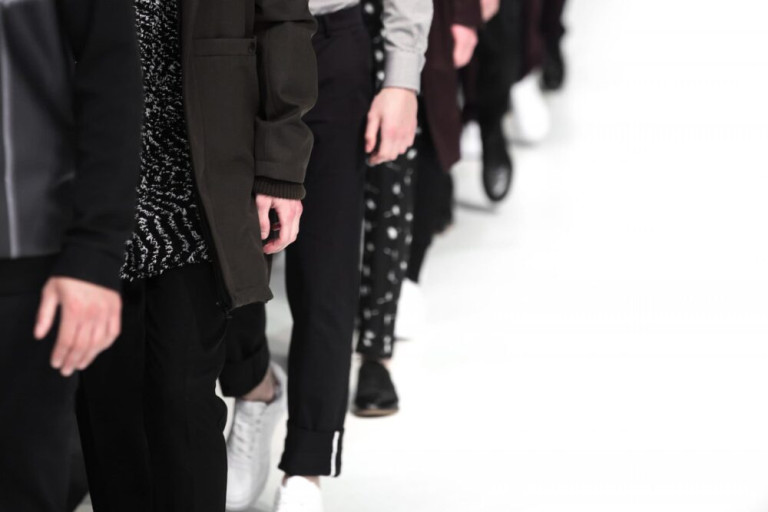 A row of people walking on a runway, dressed in stylish, modern clothing with mostly dark tones. Only their lower bodies and hands are visible against a white background.