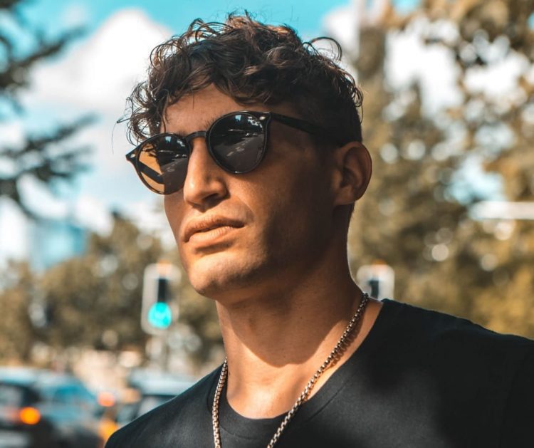 A man with curly hair wearing round sunglasses and a black shirt stands outdoors on a sunny day, with blurred trees and traffic lights in the background.