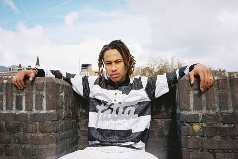 A person with medium-length dreadlocks sits on a brick wall, wearing a black and white patterned shirt with “Patta” written on it, arms stretched out on the wall, with a cloudy sky and buildings in the background.