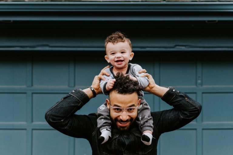 A smiling man with glasses holds a happy baby on his shoulders. The baby is holding onto the mans head, both are looking at the camera and smiling, standing in front of a blue garage door.