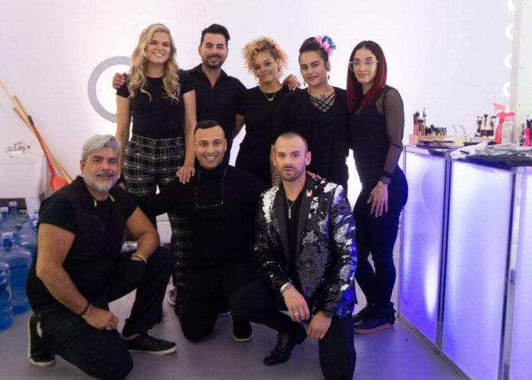 A group of eight people, dressed mostly in black, pose and smile together in a brightly lit room with makeup stations and water bottles visible in the background.