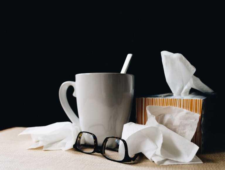 A white mug with a spoon, a box of tissues, scattered tissues, and a pair of eyeglasses on a table against a dark background.
