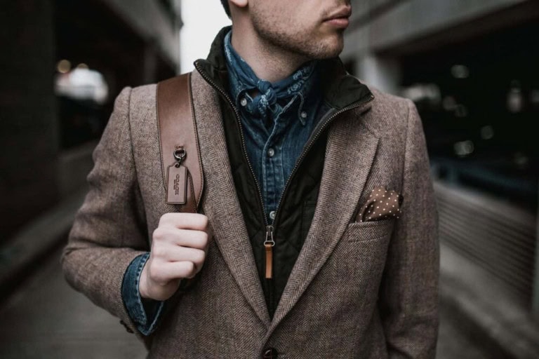 A man wearing a brown textured blazer, blue button-up shirt, and black vest walks outdoors while carrying a brown leather shoulder bag. A brown polka-dot pocket square is visible in his blazer pocket.