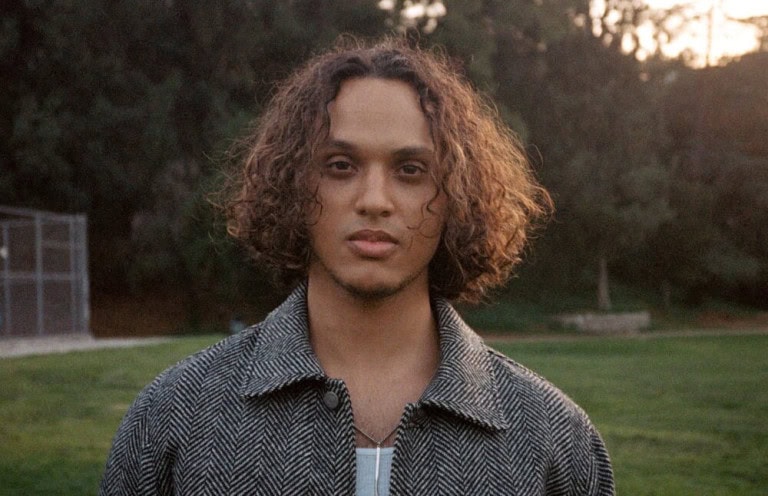 A person with curly, shoulder-length hair stands outdoors on grass, wearing a herringbone-patterned jacket over a light shirt, with trees and a fence blurred in the background.