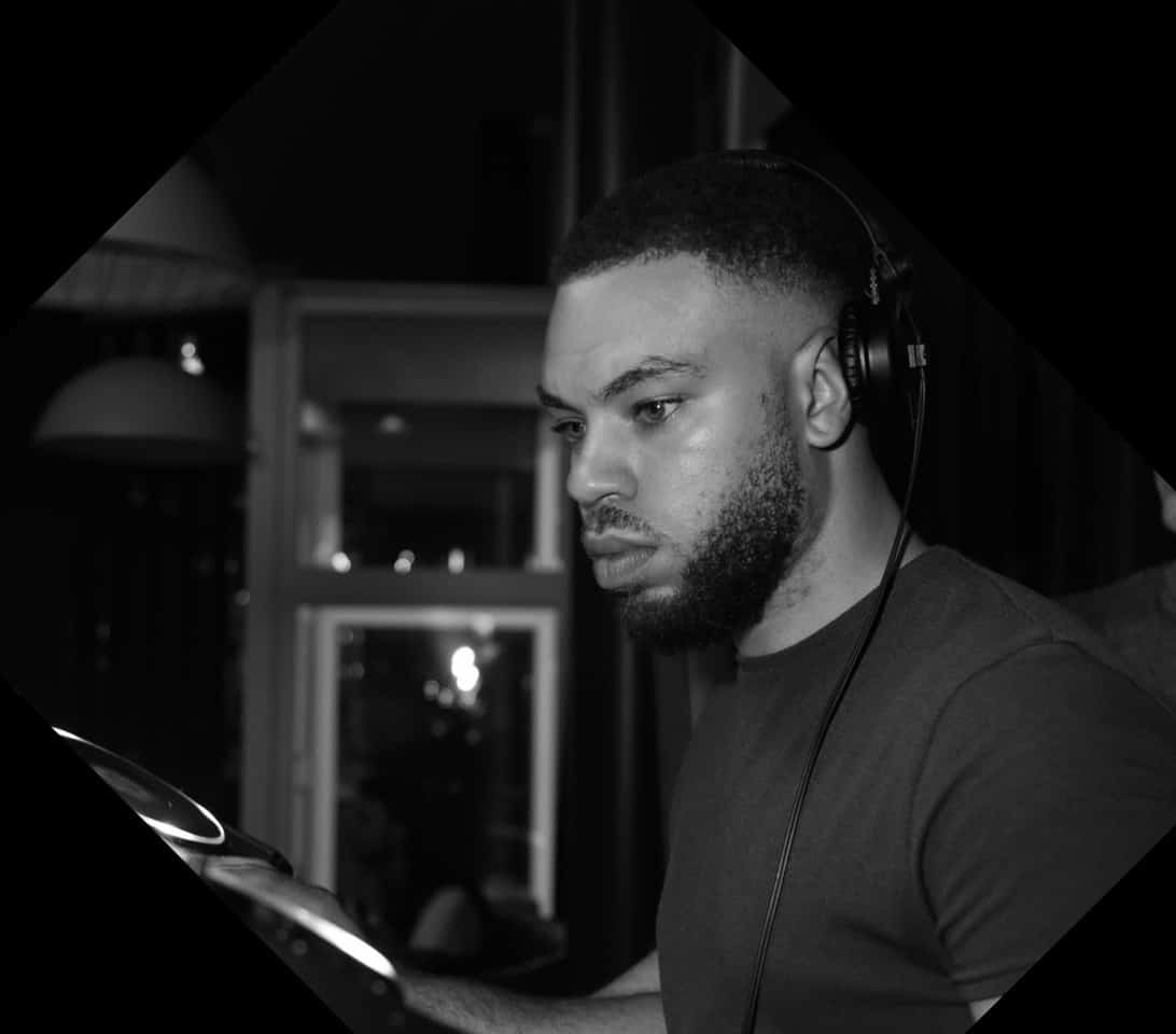 A man with headphones is focused while using DJ equipment in a dimly lit room. The photo is in black and white.