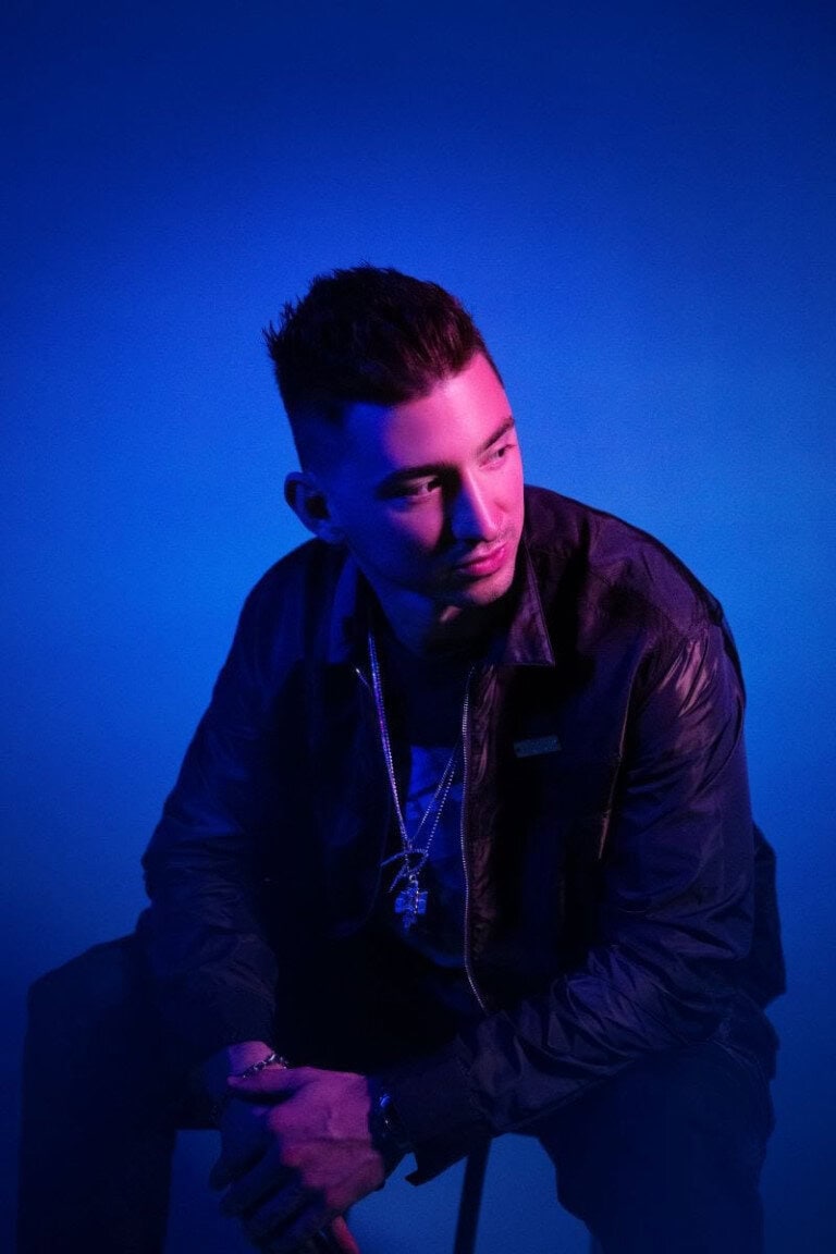 A man with short dark hair, wearing a black jacket and multiple necklaces, sits on a stool against a blue background with dramatic lighting.