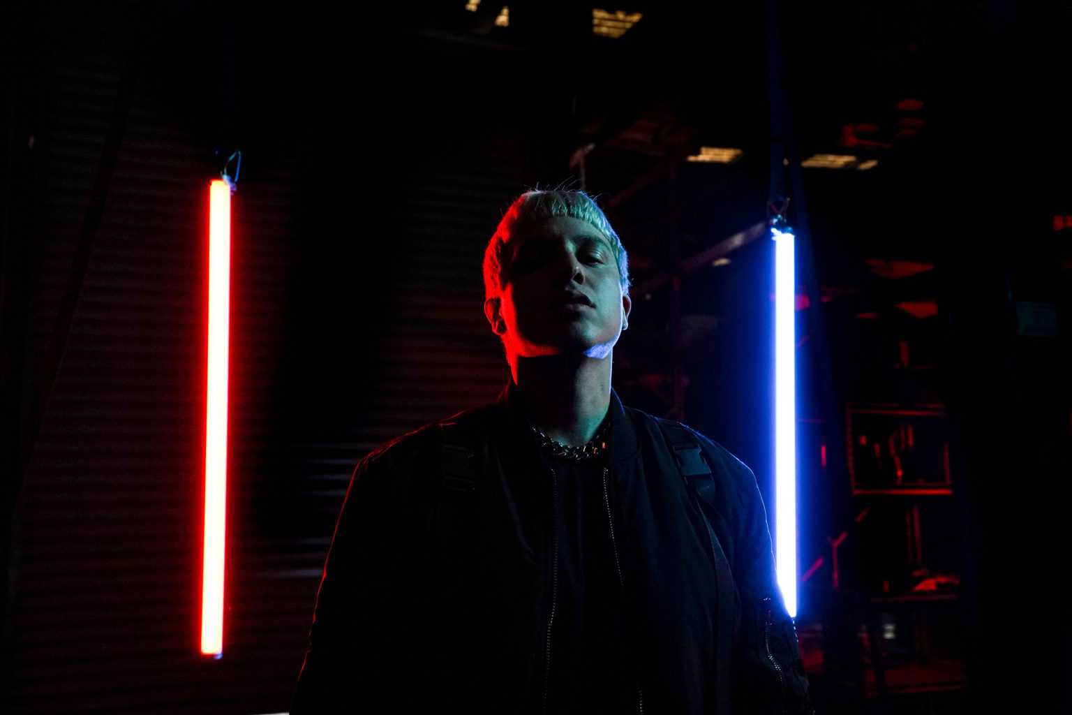 A person stands in darkness between two vertical neon lights, one red and one blue, casting dramatic shadows on their face and highlighting their silhouette.
