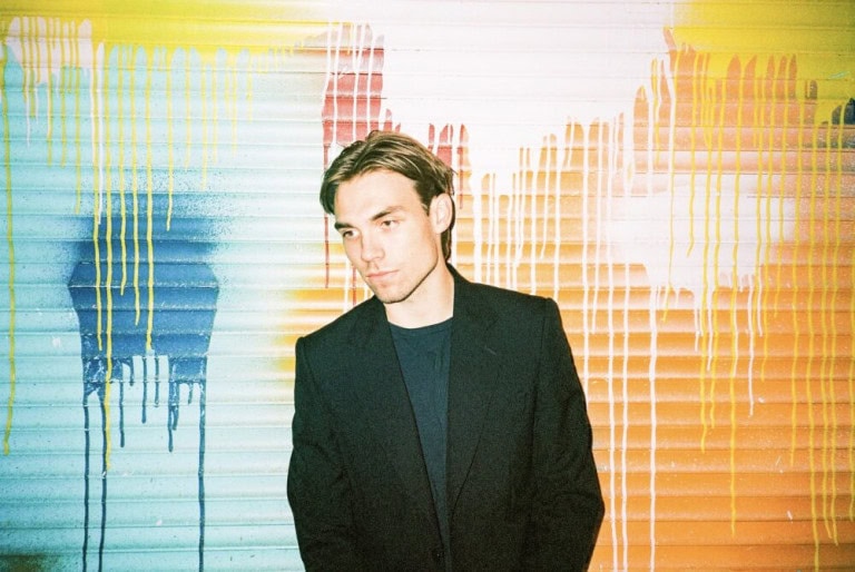 A young man in a dark blazer stands in front of a wall with colorful paint drips in yellow, blue, orange, and white. He looks slightly to the side, with a neutral expression.