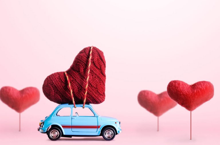 A small blue toy car carries a large red knitted heart on its roof, with two red heart-shaped decorations on sticks in the background against a soft pink backdrop.