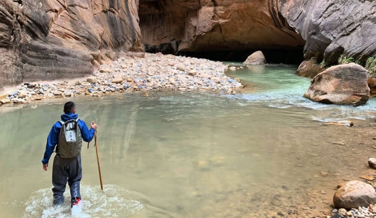 A person wearing outdoor gear and carrying a walking stick wades through a shallow river inside a narrow canyon with rocky walls and a mix of water and stones.