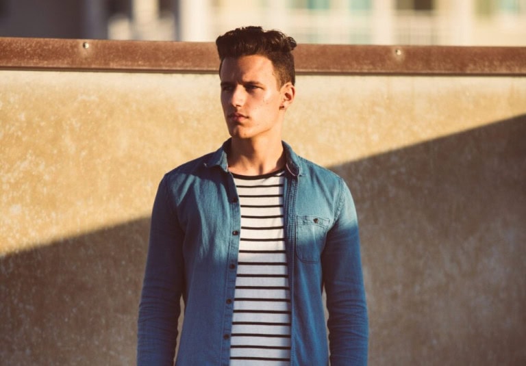 A young man with short brown hair stands outside in sunlight, wearing a blue denim shirt over a white and black striped t-shirt, with a concrete wall and blurred buildings in the background.