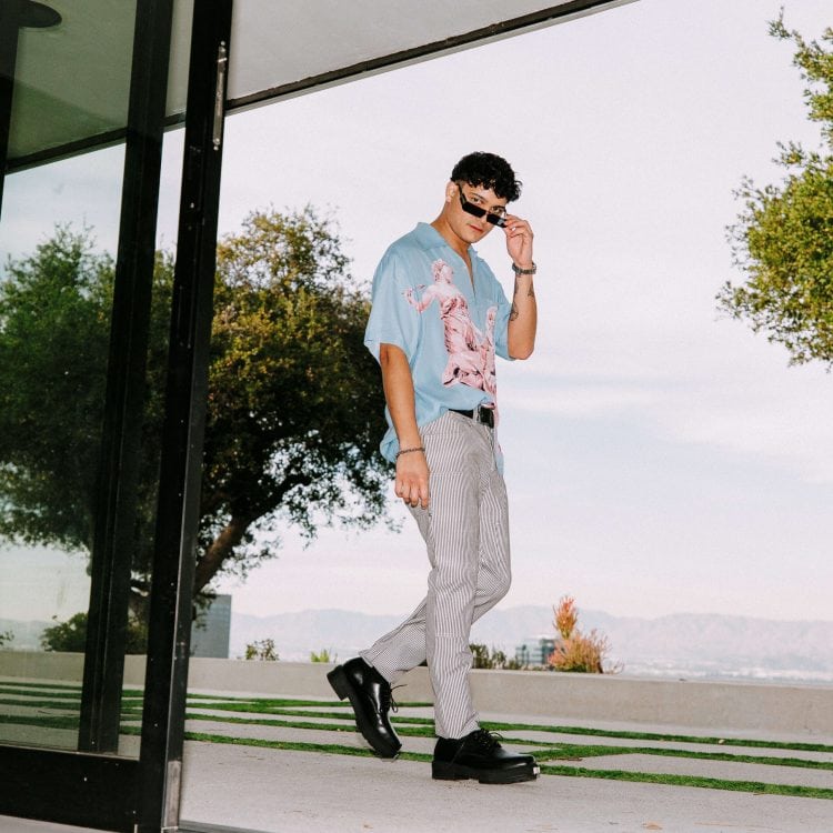 A man wearing sunglasses, a blue graphic shirt, striped pants, and black shoes walks outside near a modern building with large windows and green landscaping; trees and mountains are visible in the background.