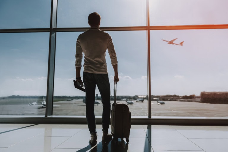 A man stands in an airport terminal holding a suitcase and a laptop, looking out large windows at an airplane taking off outside on a sunny day.
