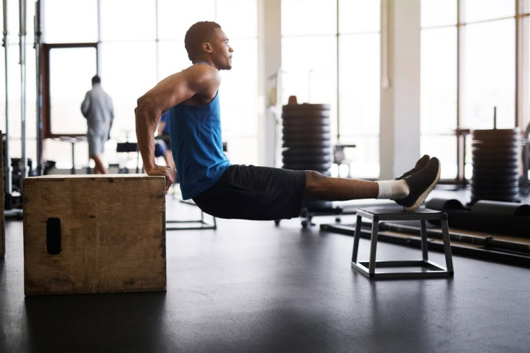 A man in athletic wear performs a tricep dip with his hands on a wooden box and his feet on a metal bench in a gym with large windows and gym equipment in the background.