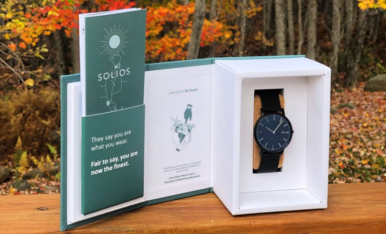 A Solios watch in a white box sits on a wooden surface outdoors, with autumn trees in the background. The open box reveals the black watch and a booklet with the Solios logo and inspirational text.