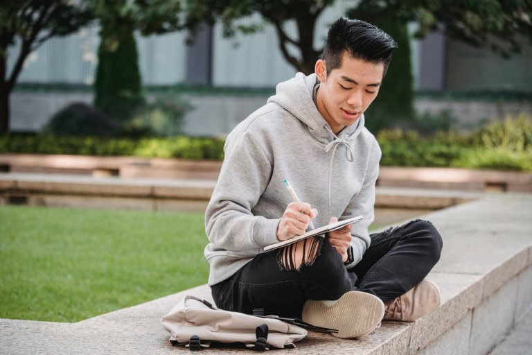A young man in a gray hoodie and ripped black jeans sits cross-legged on a stone bench outdoors, writing in a notebook with a pencil. A beige backpack is placed beside him on the bench.