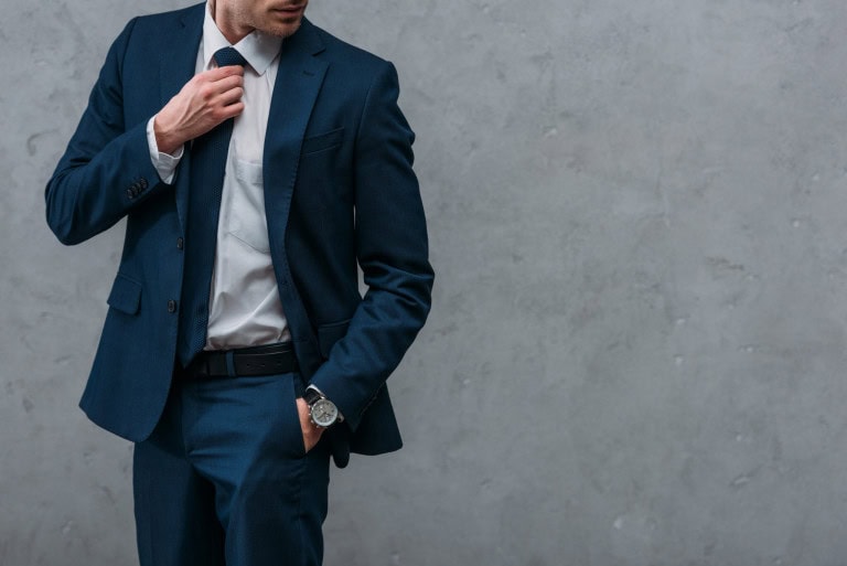 A man wearing a dark blue suit and white shirt adjusts his tie with one hand while the other hand is in his pocket. He is standing against a plain, light gray textured wall. His face is partially out of frame.