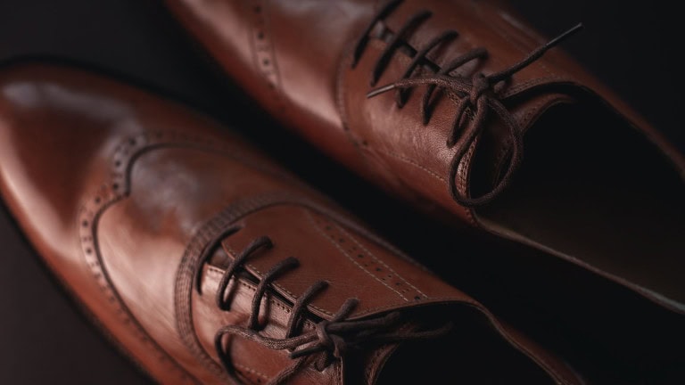 A close-up view of a pair of polished brown leather dress shoes with black laces, showing detailed stitching and brogue patterns on a dark background.