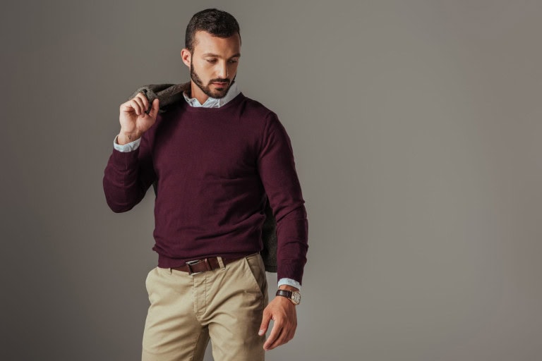 A man wearing a maroon sweater over a collared shirt and khaki pants holds a jacket over his shoulder and looks down, standing against a plain gray background.