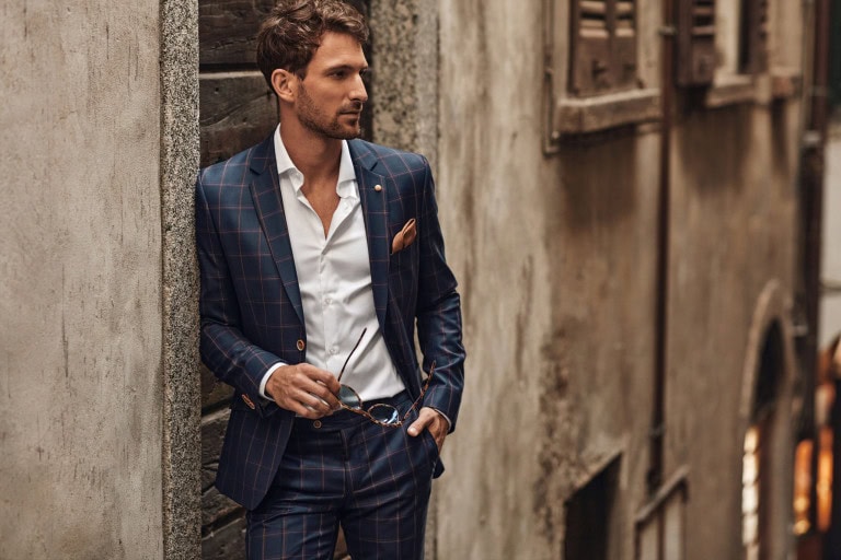 A man in a blue checked suit leans against a wall in a narrow street, holding sunglasses in one hand and looking to the side. He wears a white shirt and has a pocket square in his suit jacket.