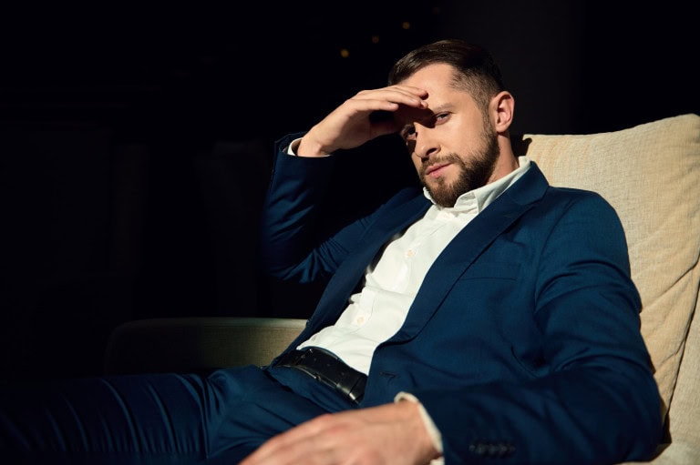 A man in a blue suit sits on a beige chair, leaning back with one arm resting and the other hand shielding his eyes from the light, looking thoughtfully into the distance against a dark background.