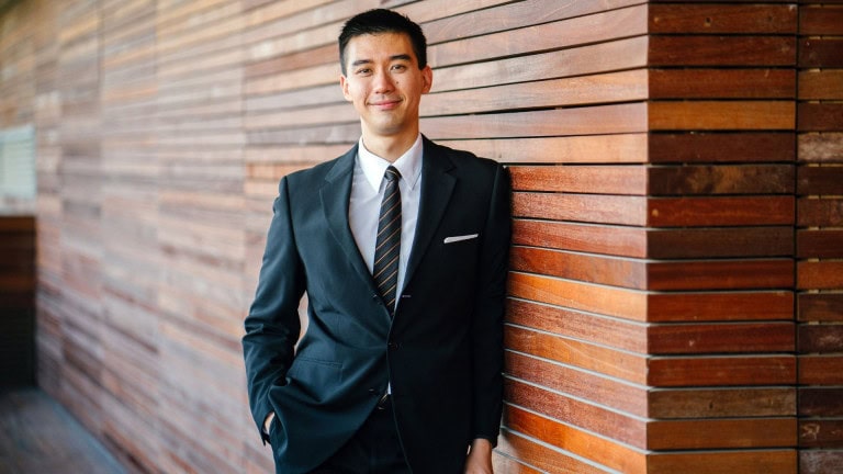 A man in a black suit and striped tie stands with one hand in his pocket, leaning against a wall made of horizontal wooden planks, smiling confidently at the camera.
