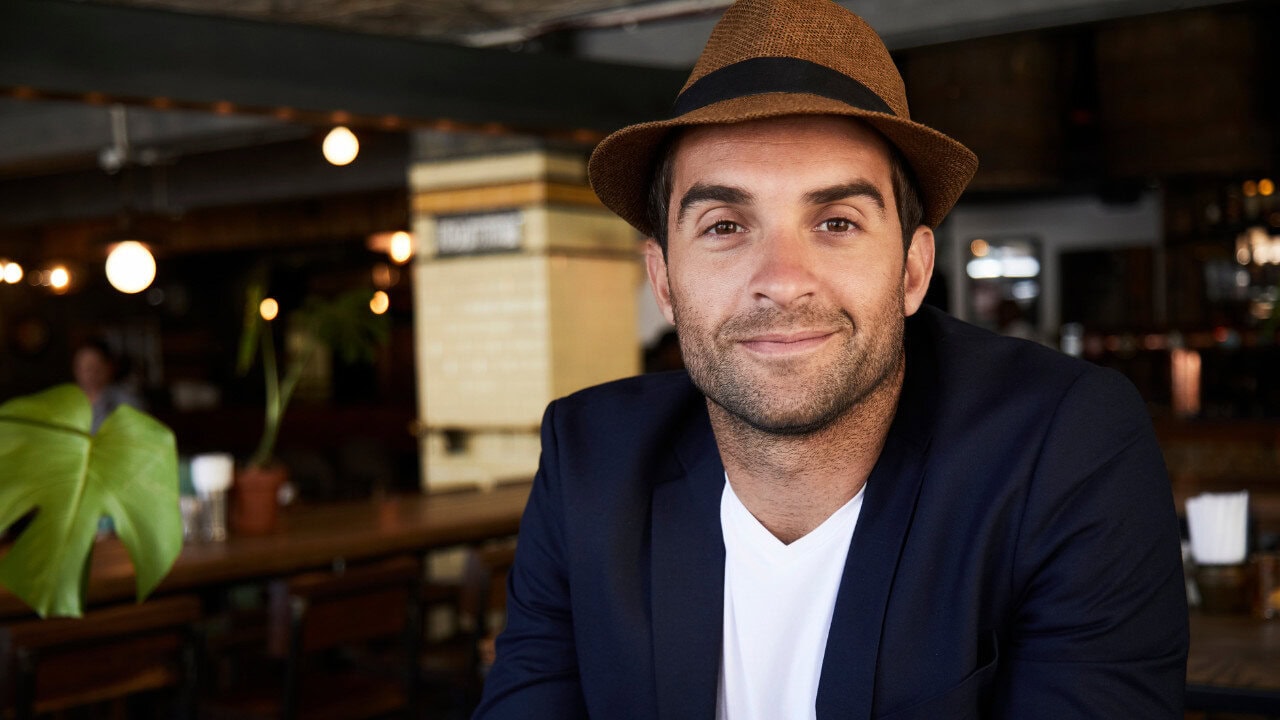 A man wearing a brown fedora, navy blazer, and white T-shirt smiles while sitting in a cozy cafe with wooden tables and warm lighting.