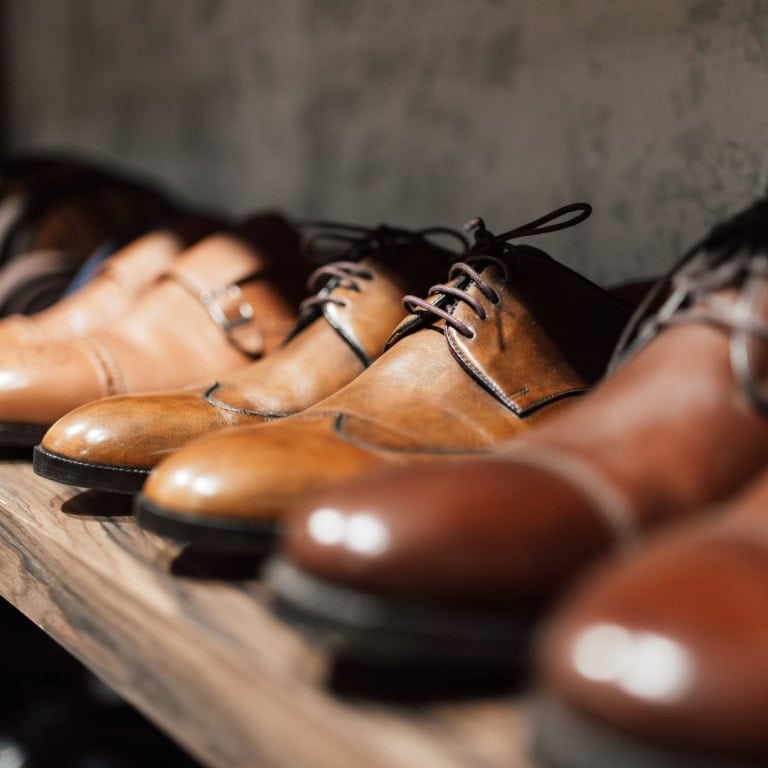 A close-up of several pairs of brown leather dress shoes neatly lined up on a wooden shelf, with the focus on the central pair and a blurred background.