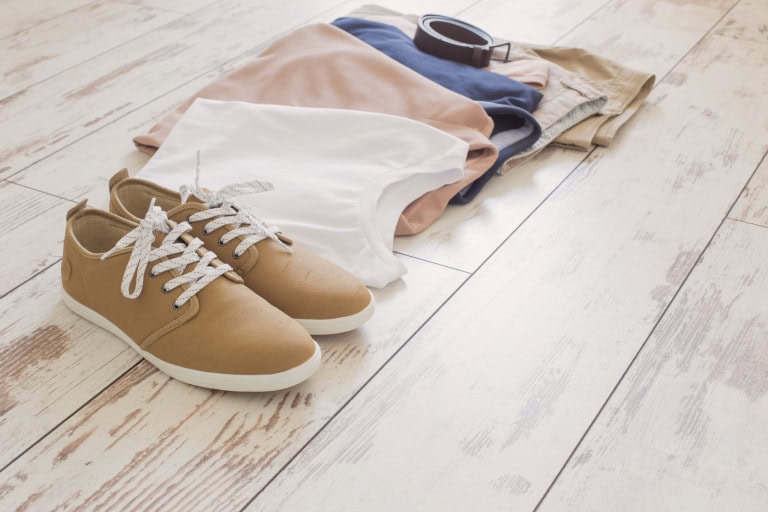 Casual outfit laid out on a wooden floor: tan sneakers, a white t-shirt, a beige t-shirt, a navy shirt, khaki shorts, and a brown belt. The clothes are neatly arranged in a row.