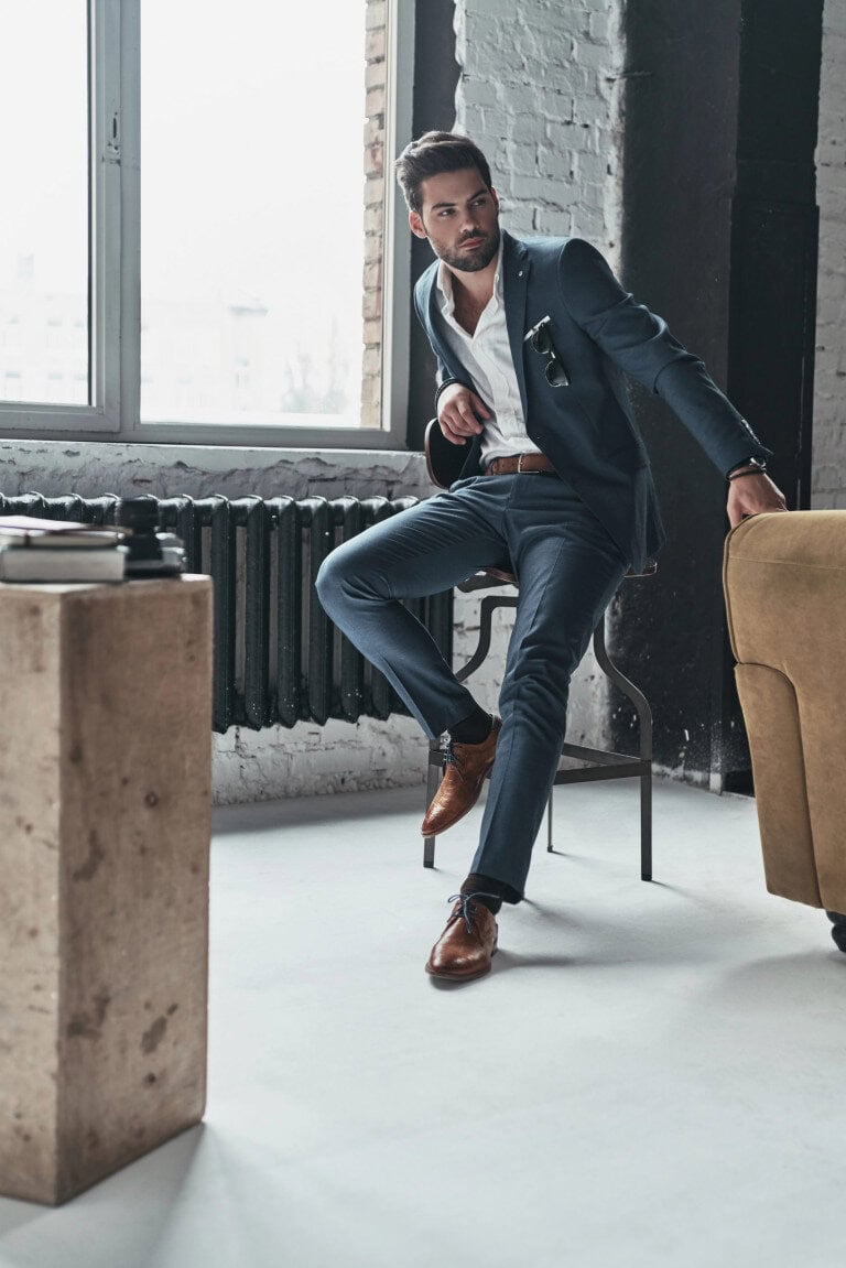 A man in a blue suit and brown shoes sits on a chair in a modern, industrial-style room, with one hand on a yellow armchair and the other adjusting his suit jacket. A window and books are visible in the background.