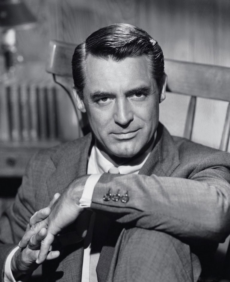 A man in a suit sits in a wooden chair, looking directly at the camera with a slight smile. His hands are folded over his knee and his dark hair is neatly combed. The photo is in black and white.