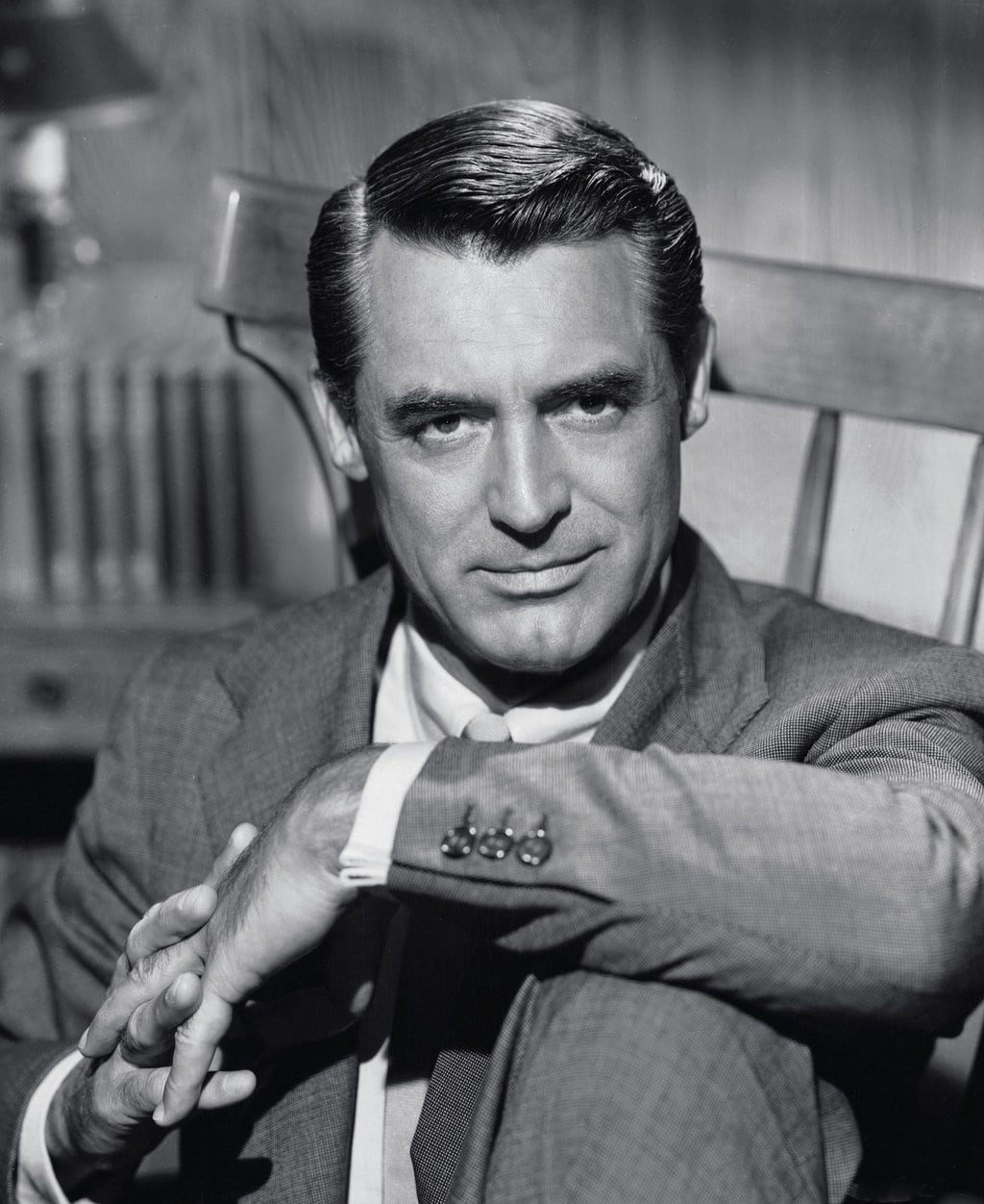 A man in a suit sits in a wooden chair, looking directly at the camera with a slight smile. His hands are folded over his knee and his dark hair is neatly combed. The photo is in black and white.