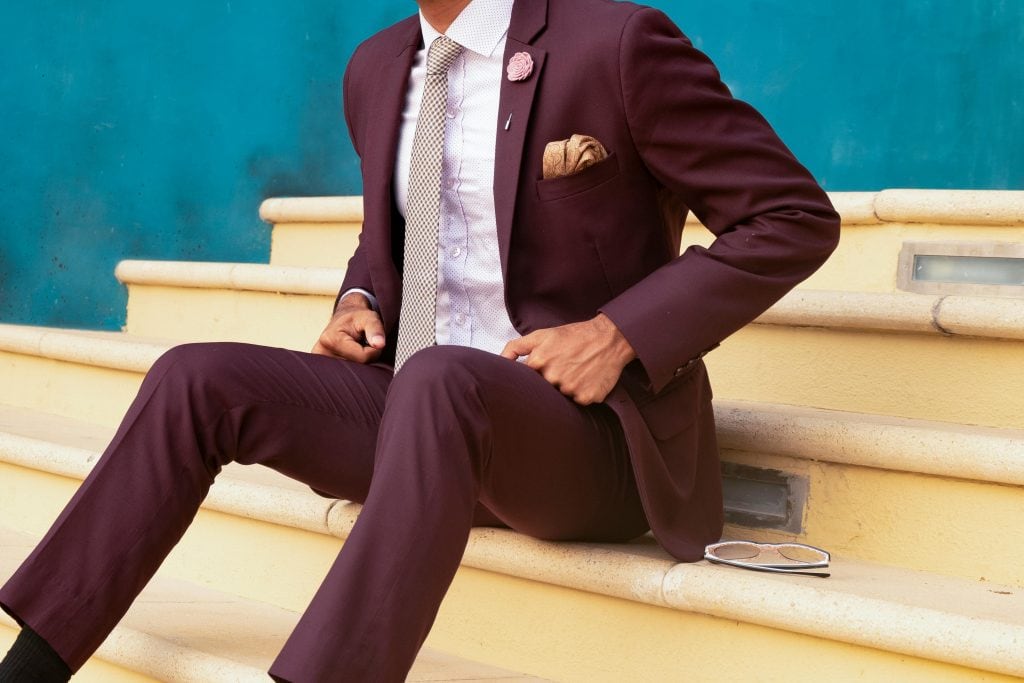 A person wearing a burgundy suit, white shirt, and light-colored tie sits on yellow steps with a turquoise wall in the background. A pair of eyeglasses lies beside them. Their face is not fully visible.