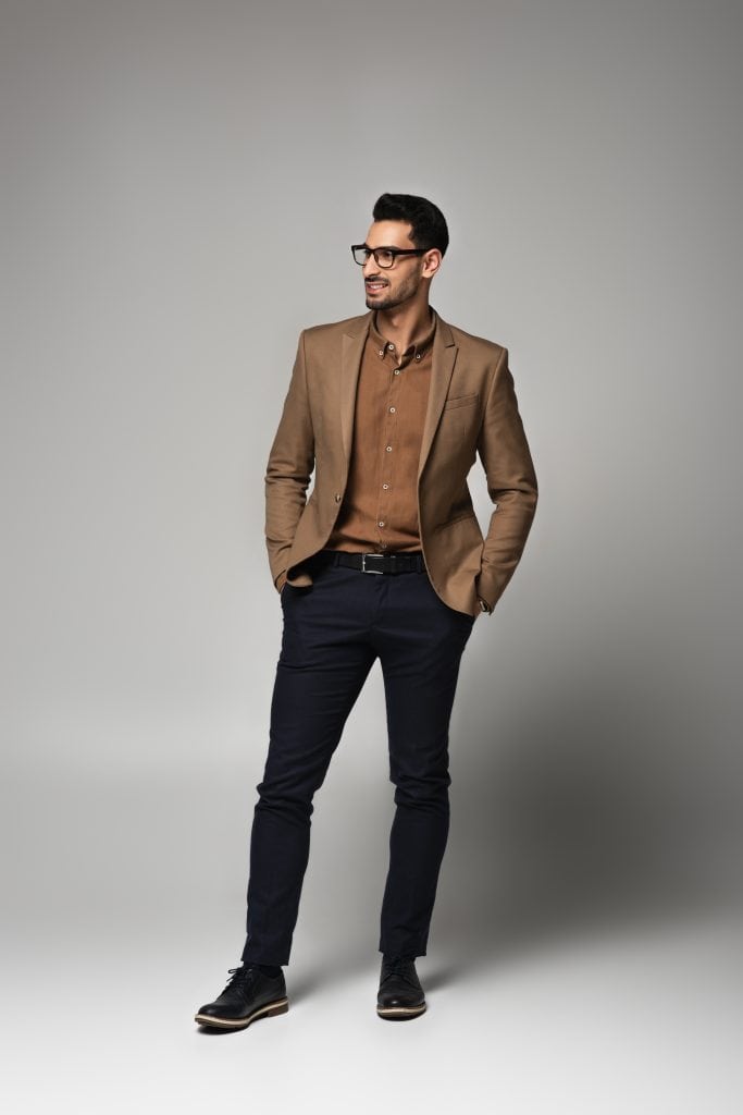 A man wearing black glasses, a brown blazer, a brown shirt, and dark pants stands with his hands in his pockets, smiling slightly, against a plain light gray background.