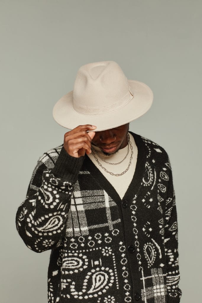 A person wearing a patterned black and white cardigan, cream shirt, chain necklaces, and a light beige hat, tilting the hat downward to partially cover their face, against a neutral background.