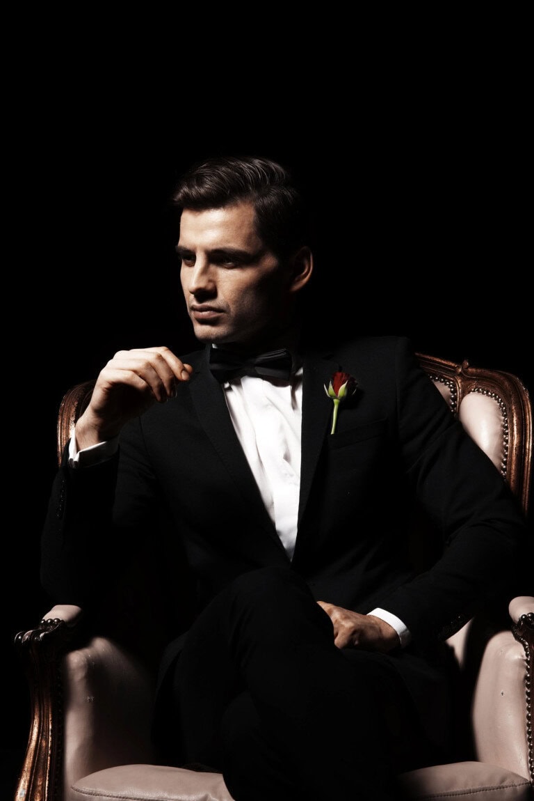 A man in a black tuxedo with a bow tie and a red rose boutonniere sits confidently on a vintage armchair against a black background, looking to the side with a composed expression.