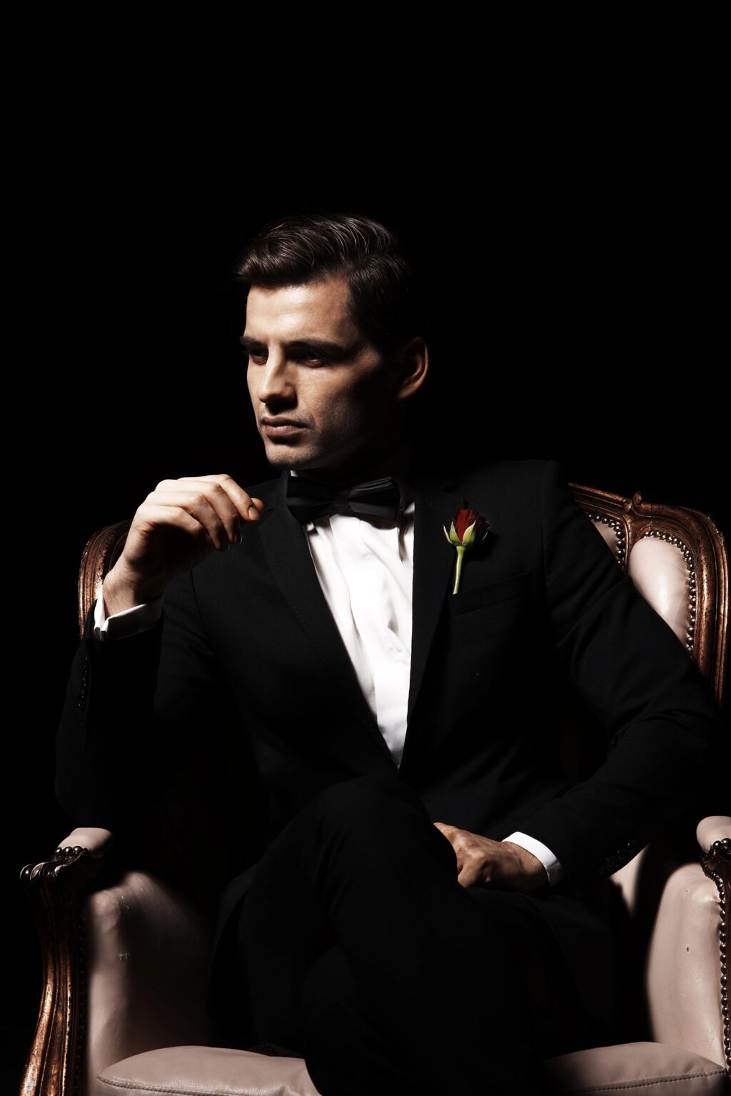A man in a black tuxedo with a bow tie and a red rose boutonniere sits confidently on a vintage armchair against a black background, looking to the side with a composed expression.