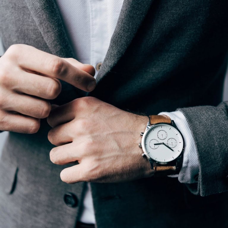 A person in a grey suit adjusts their jacket sleeve, revealing a stylish wristwatch with a brown leather strap and a white watch face. The background is out of focus.