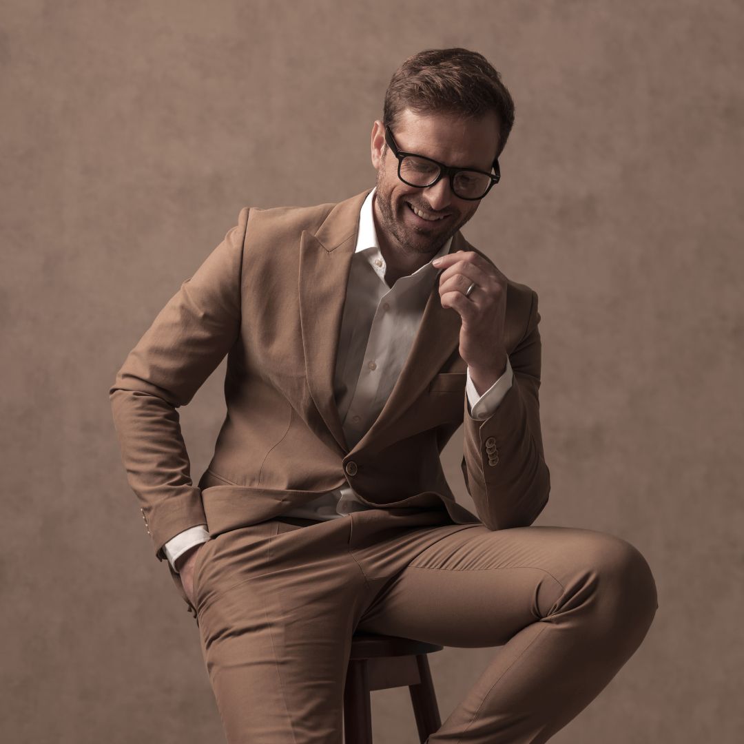 A smiling man in a tan suit and white shirt sits on a stool against a plain brown background, wearing glasses and holding his chin with one hand.