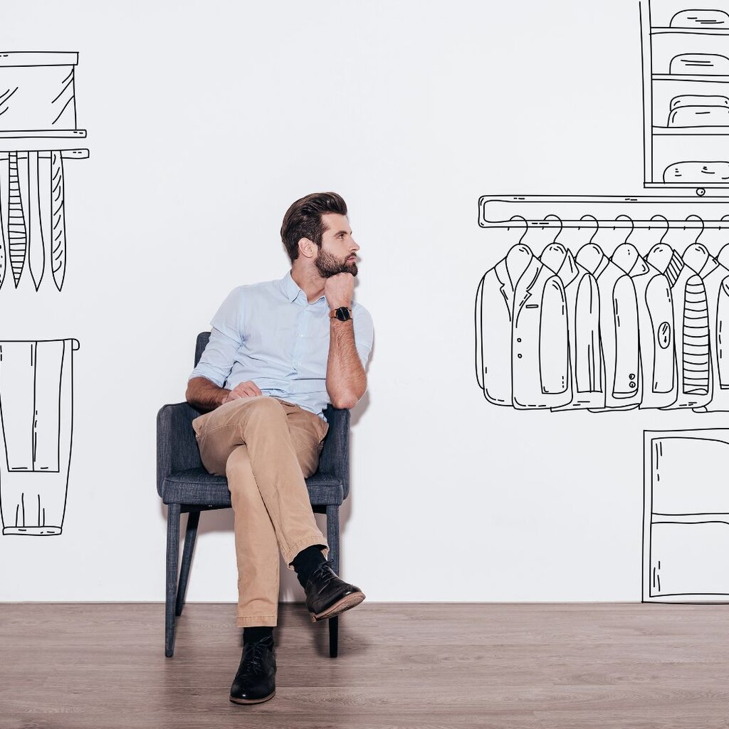 A man sits on a chair against a white wall decorated with black line drawings of a closet and hanging clothes, appearing to look thoughtfully at the illustrations.