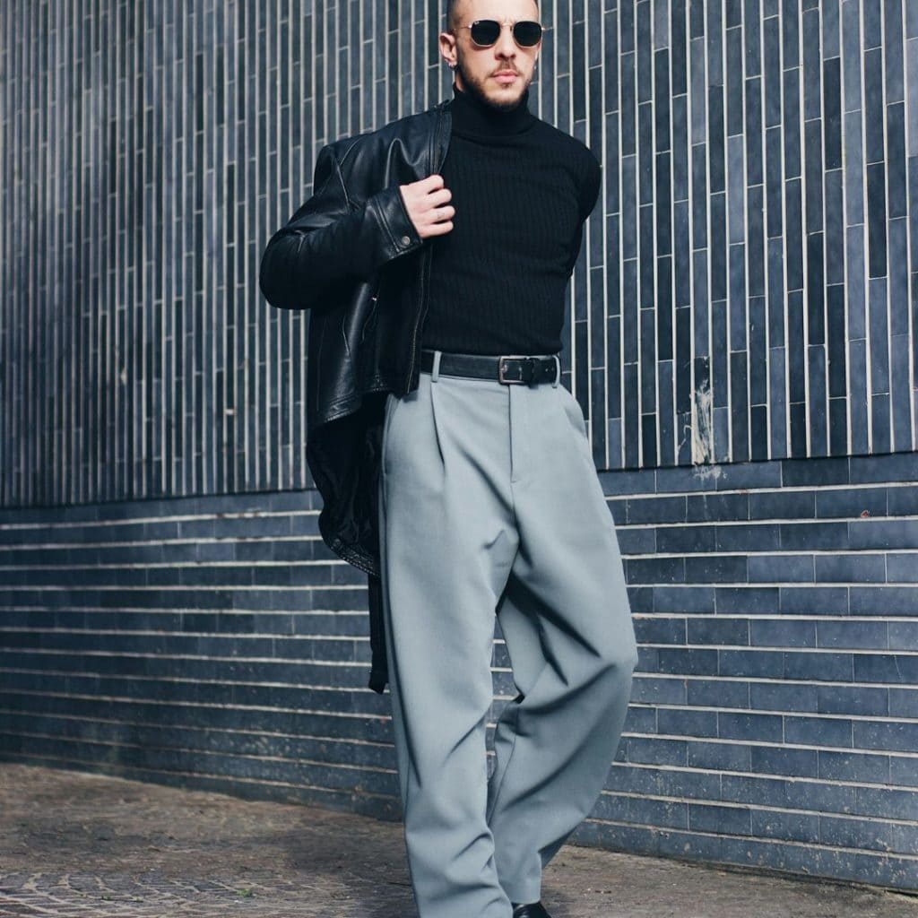 A man wearing sunglasses, a black turtleneck, a leather jacket over his shoulder, and loose-fitting gray trousers walks confidently along a sidewalk next to a modern, striped tile wall.