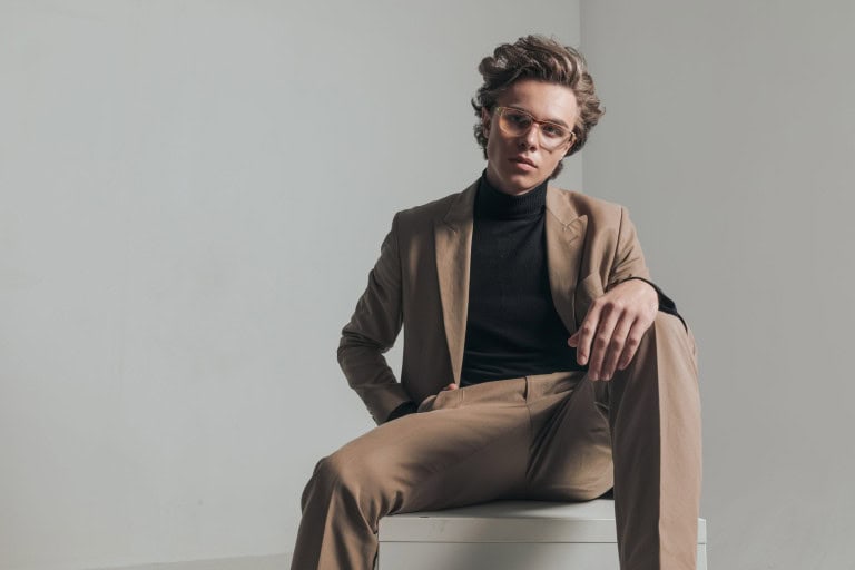 A person with wavy hair wearing glasses, a tan suit, and a black turtleneck sits confidently on a white cube against a plain, light-colored background.