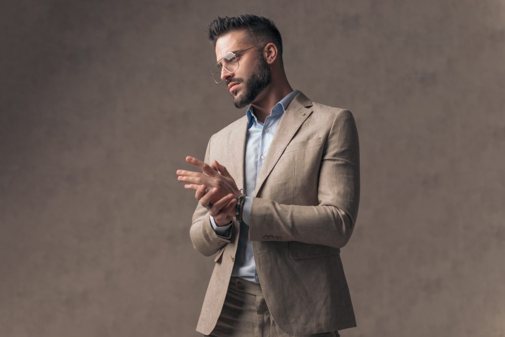 A man wearing a beige suit and light blue shirt stands against a plain, neutral background. He looks down to the side while adjusting his cuff, with a thoughtful expression on his face.