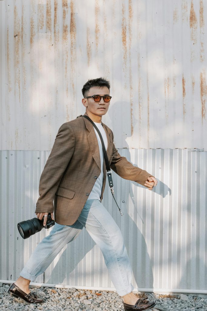 A man wearing sunglasses, a brown blazer, and light jeans walks on gravel while holding a camera. He is passing in front of a corrugated metal wall with rusty streaks.