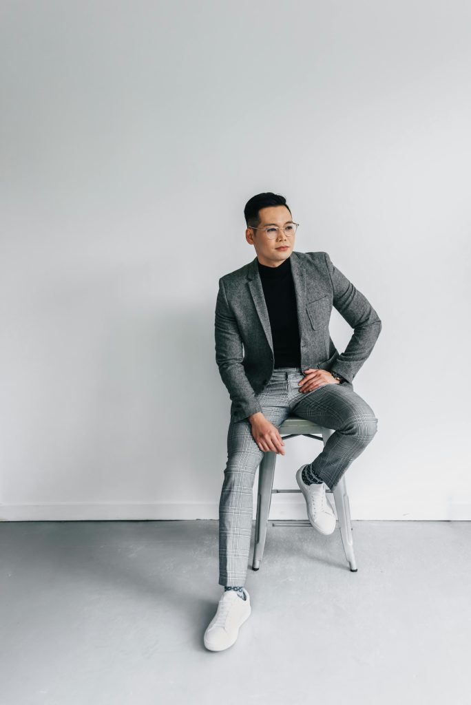 A person wearing a gray suit, black shirt, and white sneakers sits on a stool against a plain white wall, looking confidently ahead with one hand on their hip.