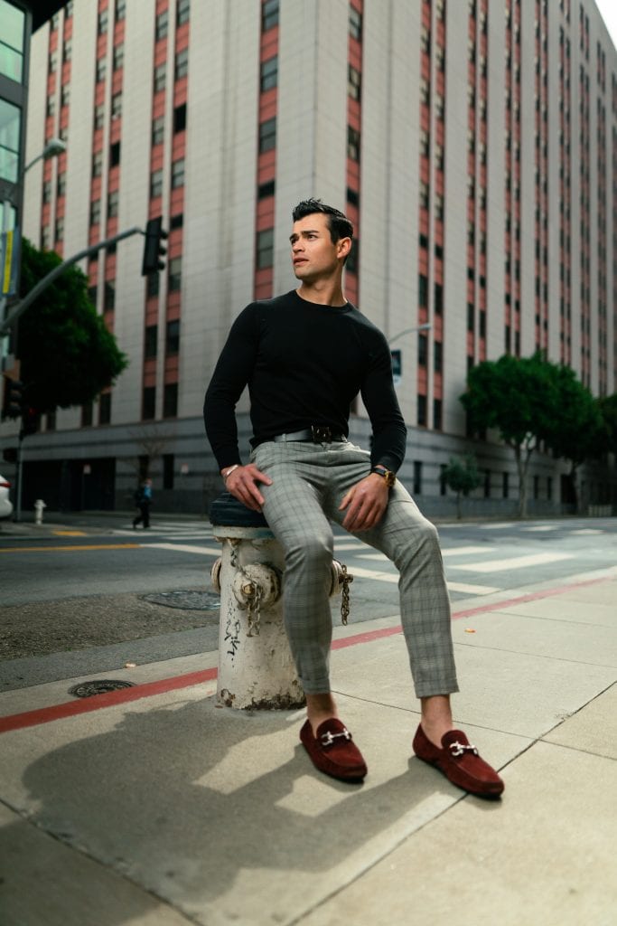 A man wearing a black long-sleeve shirt, gray plaid pants, and brown loafers sits on a fire hydrant on a city sidewalk, with a tall building and trees in the background.