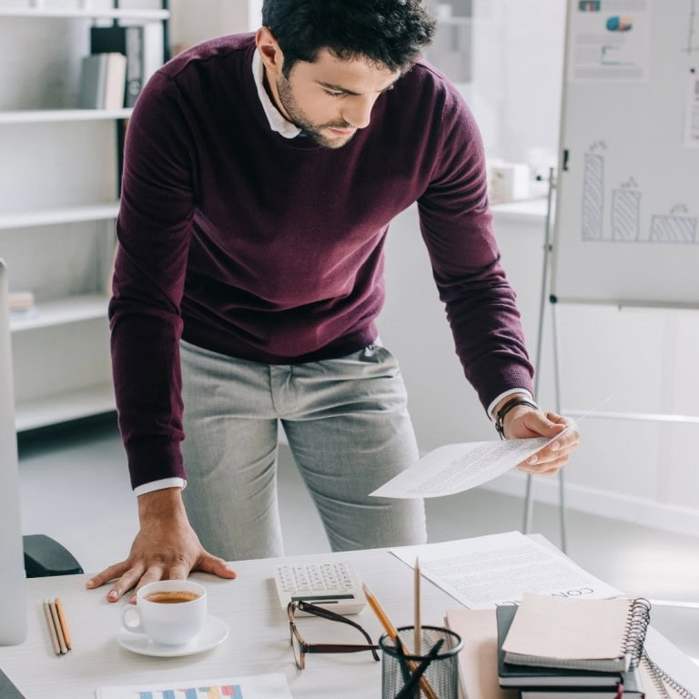 A man in a maroon sweater leans over a desk, reading a document. The desk holds papers, a cup of coffee, and office supplies. In the background, there is a whiteboard with charts and shelves.