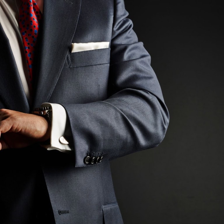 A man in a dark suit, white shirt, and red patterned tie adjusts his watch. Only his torso and arms are visible against a dark background. A white pocket square is tucked into his jacket pocket.