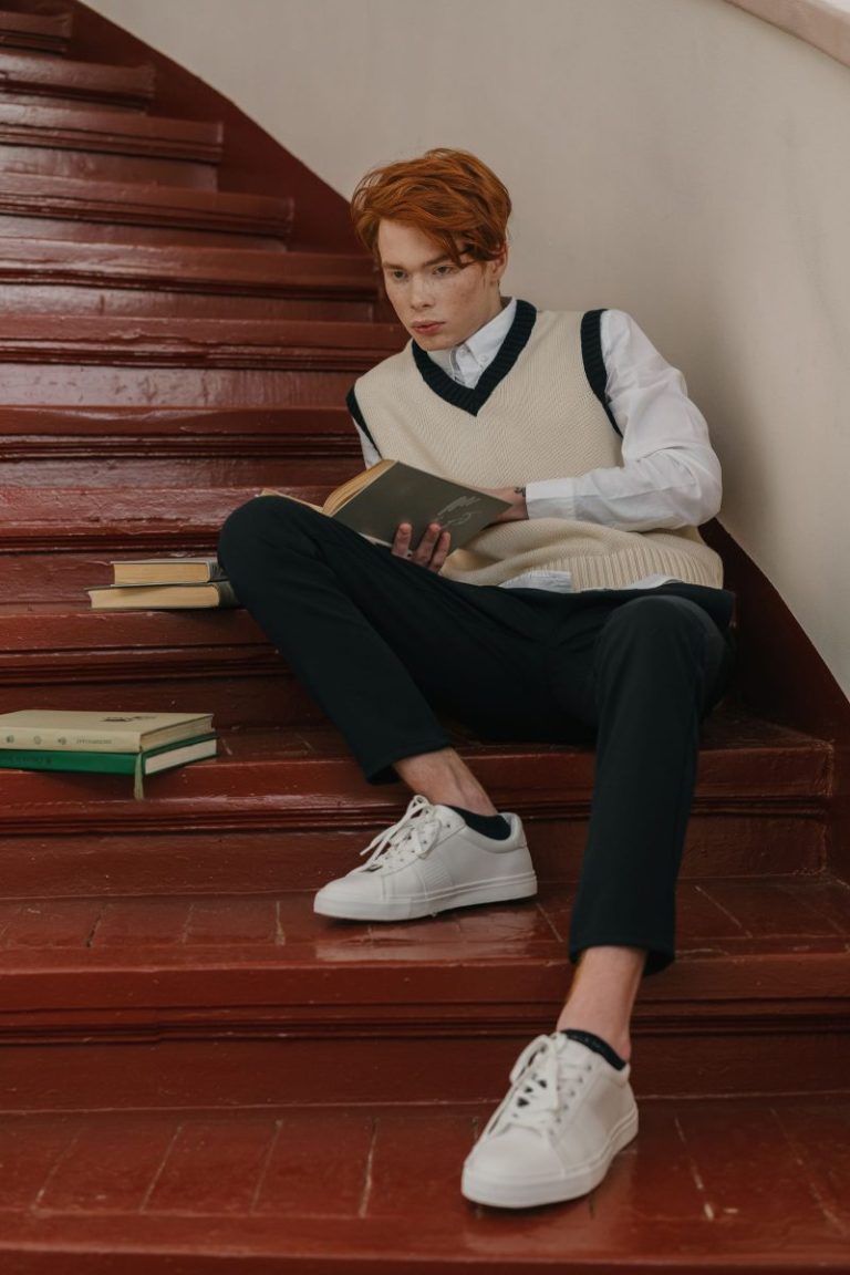 Young man in a sweater vest reading a book on steps, Ivy style