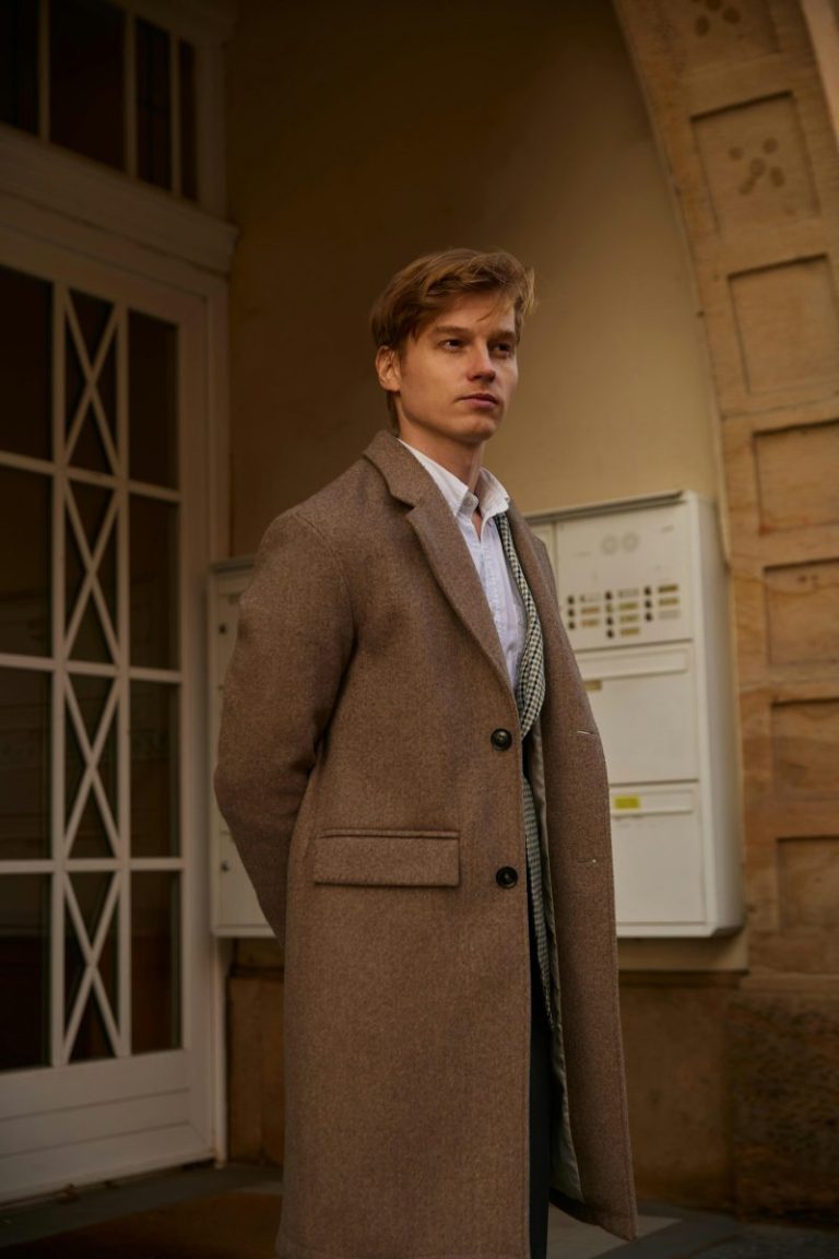 Man in a camel overcoat standing under a stone archway, quiet luxury style